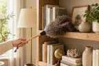 Person using a duster to clean a wooden shelf with books and decorative items.