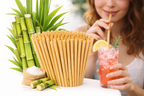 Bowl of sugar cane straws with a woman drinking a pink iced drink, surrounded by greenery.