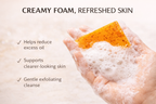 Hand with foamy skin, orange scrubber, and text about skincare benefits on a light background