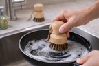 Person cleaning a frying pan with a wooden scrubber in a kitchen sink.